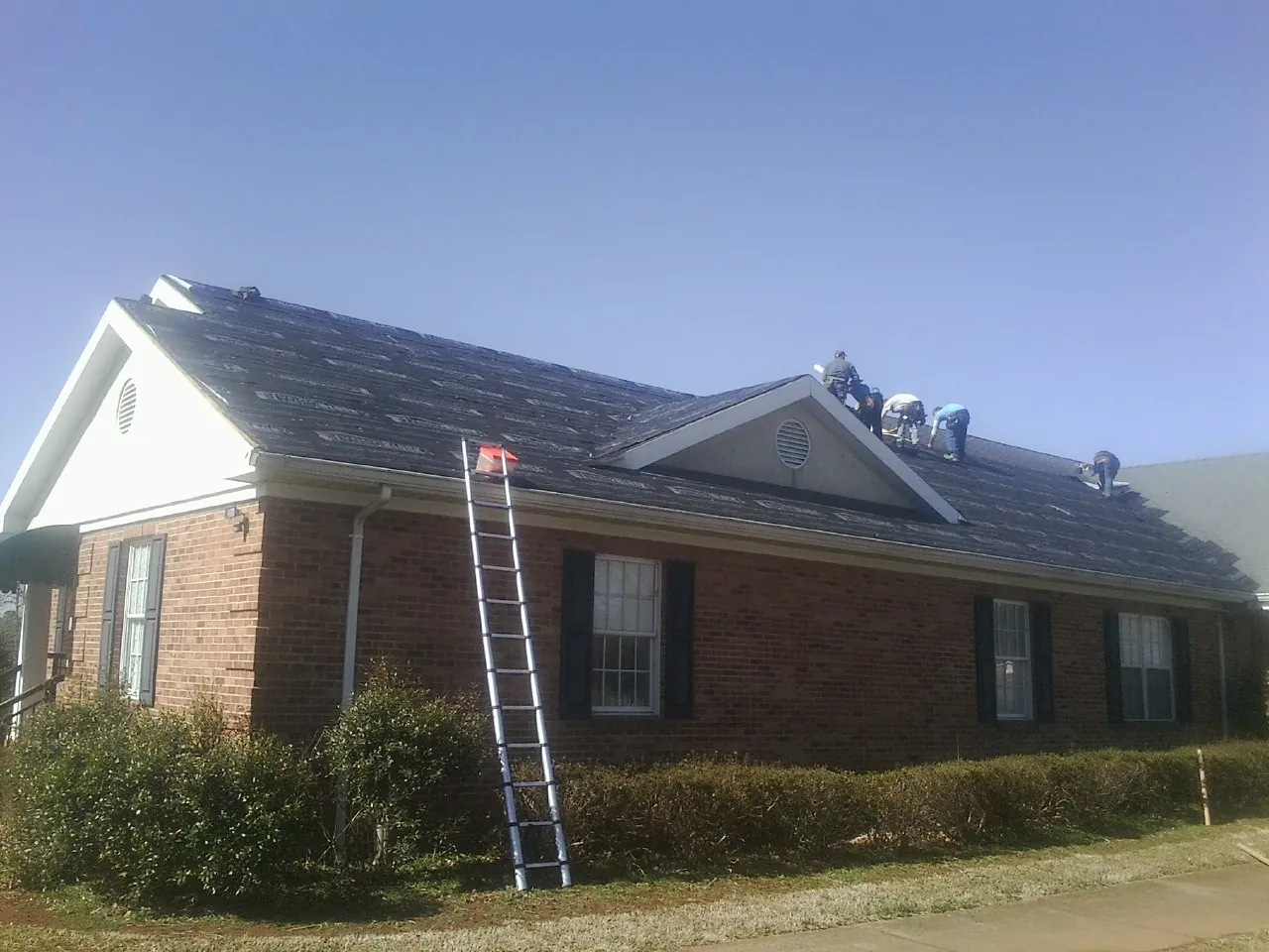 Crew tearing shingles off a brick ranch roof with a ladder set — Metal Roof Repair work by our crew in Whitesville